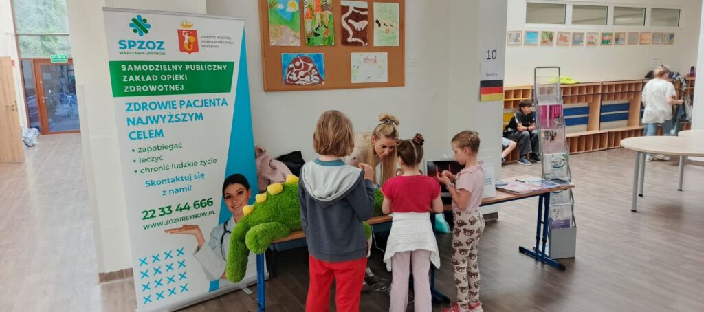 Pani prowadząca zajęcia w Szpitalu Pluszowego Aligatora pokazuje dzieciom ze Szkoły Podstawowej nr 81, jak badać pluszowego pacjenta. Dzieci z zainteresowaniem obserwują i uczestniczą w edukacyjnej zabawie, poznając podstawy opieki zdrowotnej w przyjaznej atmosferze.