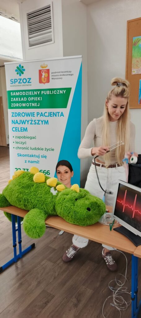 Pani prowadząca zajęcia w Szpitalu Pluszowego Aligatora pokazuje dzieciom ze Szkoły Podstawowej nr 81, jak badać pluszowego pacjenta. Dzieci z zainteresowaniem obserwują i uczestniczą w edukacyjnej zabawie, poznając podstawy opieki zdrowotnej w przyjaznej atmosferze.
