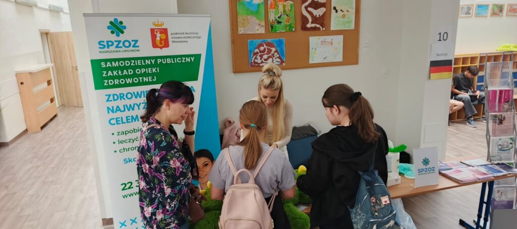 Pani prowadząca zajęcia w Szpitalu Pluszowego Aligatora pokazuje dzieciom ze Szkoły Podstawowej nr 81, jak badać pluszowego pacjenta. Dzieci z zainteresowaniem obserwują i uczestniczą w edukacyjnej zabawie, poznając podstawy opieki zdrowotnej w przyjaznej atmosferze.